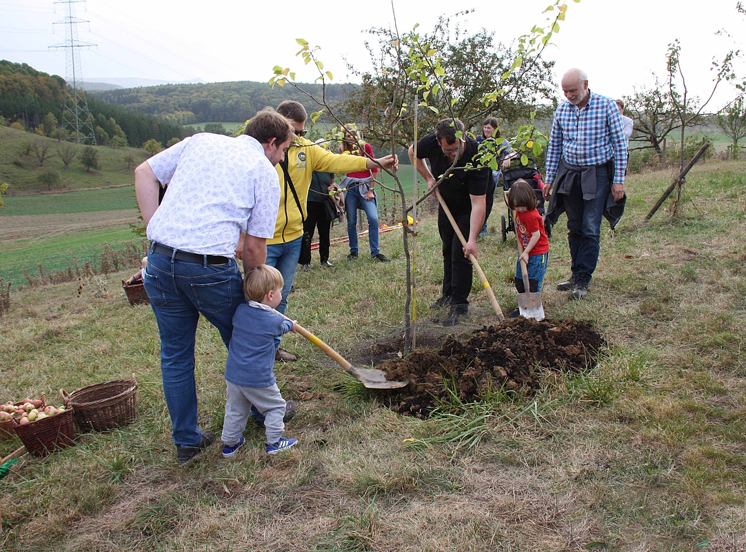 Eltern und Kinder schaufeln Erde auf das Loch in dem der neue geplanze Baum steht