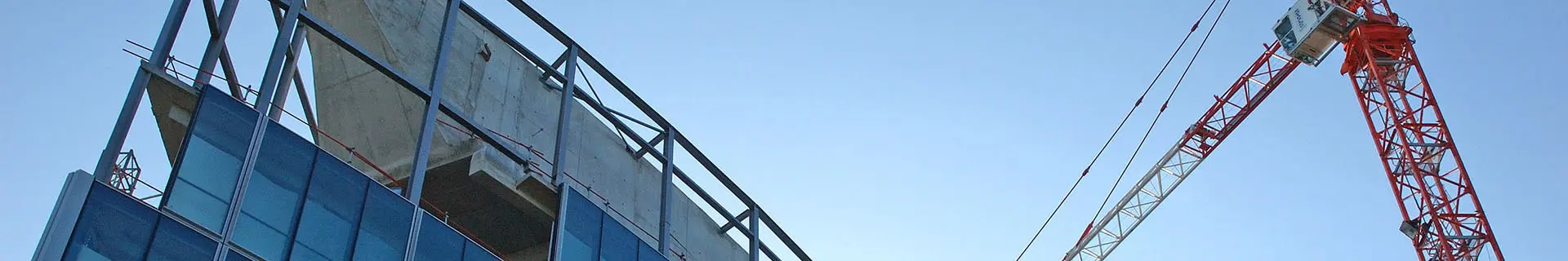 Das Bild zeigt eine Baustelle mit einem teilweise errichteten Gebäude und einem großen rot-weißen Baukran. Die unteren Stockwerke des Gebäudes sind bereits mit Glasfenstern versehen, die oberen Stockwerke sind noch im Rohbau mit sichtbarem Beton und Stahlgerüst. Der Kran ragt hoch über das Gebäude und deutet auf aktive Bauarbeiten hin. 