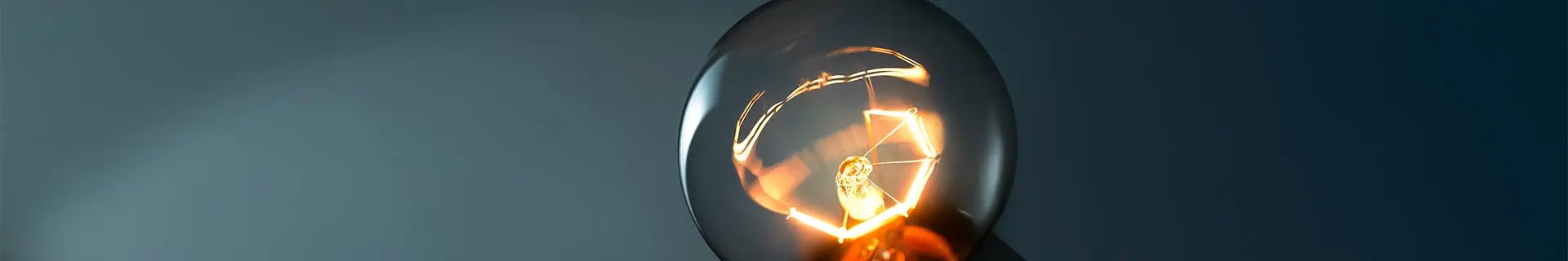 Das Bild zeigt eine Nahaufnahme einer leuchtenden Glühbirne. Im Mittelpunkt steht der glühende Glühfaden im Inneren der Birne, der ein warmes, orangefarbenes Licht ausstrahlt. Der Hintergrund ist dunkel, wodurch die Helligkeit und die feinen Details der Filamentstruktur im Glas besonders hervorgehoben werden.