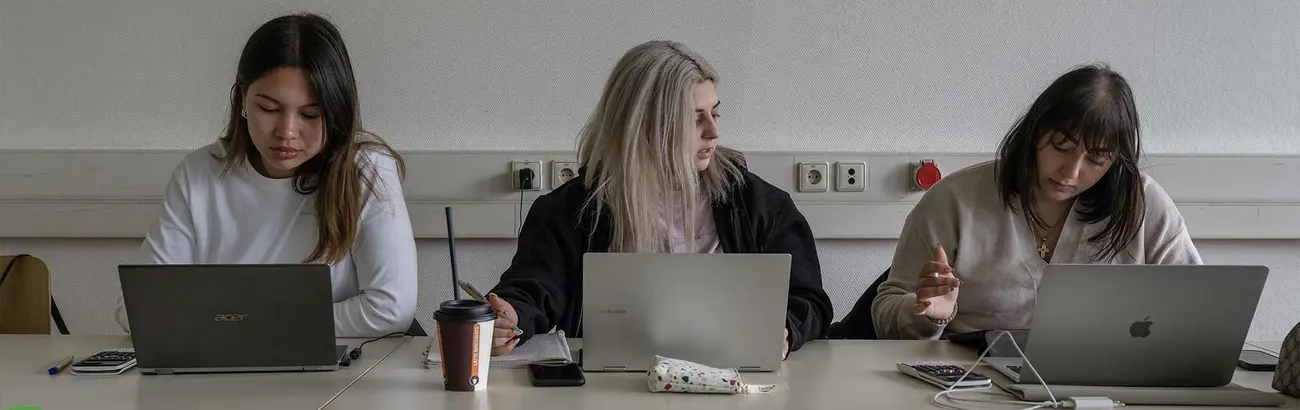 Das Bild zeigt drei Studentinnen an Laptops in einer Arbeitssituation.