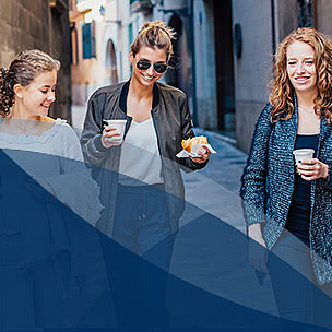 Drei Frauen in schicker Kleidung, teils mit Sonnenbrille gehen mit Kaffeebechern in der Hand eine Altstadt-Gasse entlang