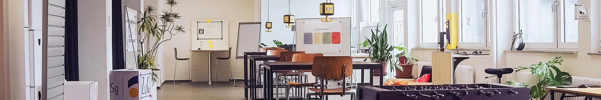 Das Bild zeigt einen modernen, gut beleuchteten Raum mit großen Fenstern auf der rechten Seite. In der Mitte stehen mehrere Tische und Stühle, darüber hängen Lampen. Auf der linken Seite befinden sich Spinde und eine große Pflanze. Im Hintergrund sieht man ein Whiteboard mit Text und Diagrammen sowie einen weiteren Tisch mit Stühlen. Im Vordergrund rechts ist ein Kickertisch zu erkennen.