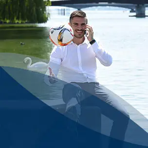 Studierender mit einem weißen Hemd sitzt auf einer Mauer vor einem See und spielt einen Fußball in die Luft