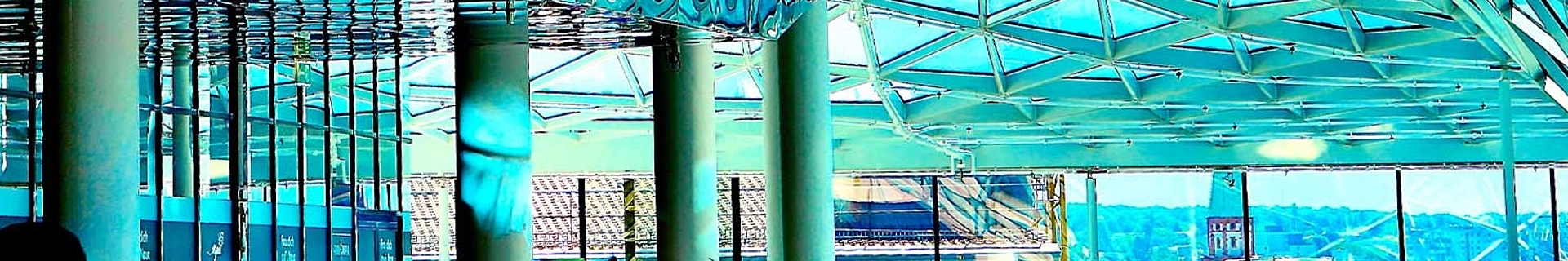 Innenraum eines modernen Gebäudes mit geometrischem Glasdach, reflektierenden Deckenpaneelen und hohen Säulen, erfüllt von natürlichem Licht und Besuchern.