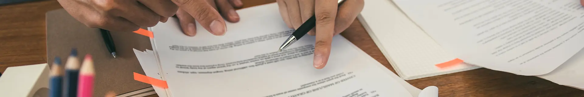 Das Bild zeigt eine Nahaufnahme von Händen, die mit Dokumenten auf einem Holztisch arbeiten. Eine Hand hält einen Stift und zeigt auf einen Text auf einem Blatt Papier, während die andere Hand ein weiteres Dokument hält oder anpasst. Auf dem Tisch liegen mehrere Blätter mit gedrucktem Text, einige davon mit orangefarbenen Markierungen. Im Hintergrund sind auch Buntstifte zu sehen. Die Szene wirkt wie eine Arbeitssituation oder ein Lernumfeld, jedoch sind keine spezifischen Aufgaben oder mathematischen Inhalte erkennbar.
