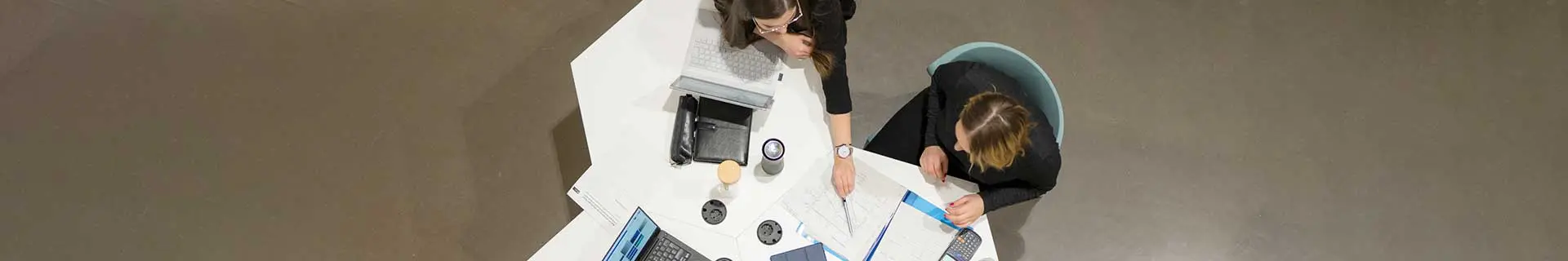 Das Bild zeigt eine Draufsicht auf zwei Personen, die an einem weißen Tisch sitzen. Eine Person arbeitet an einem Laptop, während die andere mit einem Stift auf ein Dokument zeigt. Auf dem Tisch befinden sich verschiedene Gegenstände: ein weiterer Laptop, Notizbücher, Papiere und Tassen. Es sind keine mathematischen Aufgaben oder Hausaufgaben erkennbar – die Szene wirkt wie ein Arbeits- oder Besprechungsumfeld.