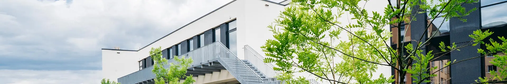 Das Bild zeigt ein modernes Gebäude mit weißer Fassade und großen Fenstern. Eine Außentreppe aus Metall führt zum oberen Stockwerk. Das Gebäude ist von grünen Bäumen umgeben, und der Himmel darüber ist bewölkt.