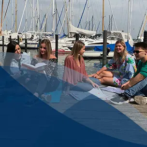 Studierende lernen und unterhalten sich auf einem Steg vor Booten bei sonnigem Wetter