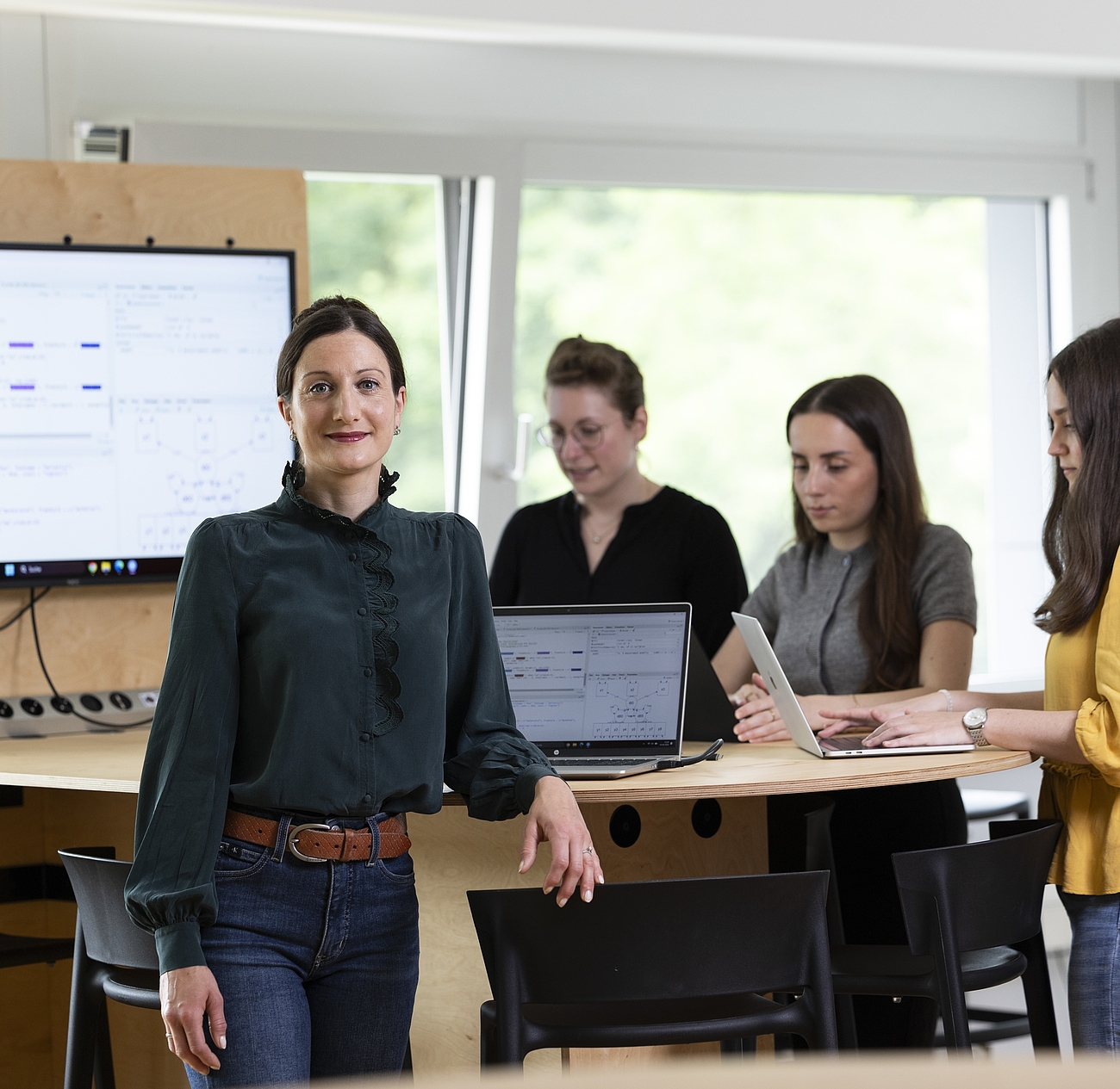 Professorin und drei Studentinnen an einem Stehtisch mit Laptops und großem Bildschirm im Hintergrund