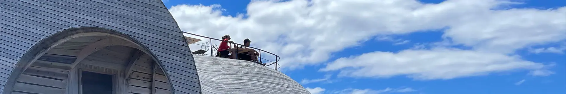 Das Bild zeigt eine geschwungene Holzstruktur mit einem bogenförmigen Fenster auf der linken Seite. Auf dem oberen Bereich der Struktur befindet sich ein Geländer, hinter dem zwei Personen sitzen und offenbar die Aussicht genießen. Der Himmel ist klar mit einigen vereinzelten Wolken.