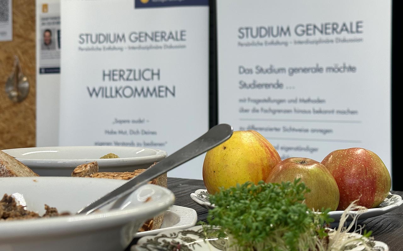 Gemüse und Obst im Vordergrund auf einem Tisch, im Hintergrund das Plakat des Studium generale