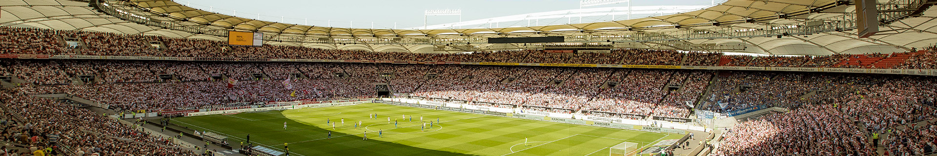 Das Bild zeigt ein großes, modernes Fußballstadion, das mit Zuschauern gefüllt ist. Die Architektur umfasst ein teilweise überdachtes Dach, und das Spielfeld ist grün und gepflegt. Auf dem Feld sind Spieler zu sehen, die gerade ein Spiel austragen. Weitere Details: Die Tribünen sind voll besetzt und sorgen für eine lebendige Atmosphäre. Entlang des Spielfeldrands befinden sich Werbebanner. Es sind verschiedene Sitzbereiche erkennbar, darunter auch Abschnitte für unterschiedliche Fangruppen.
