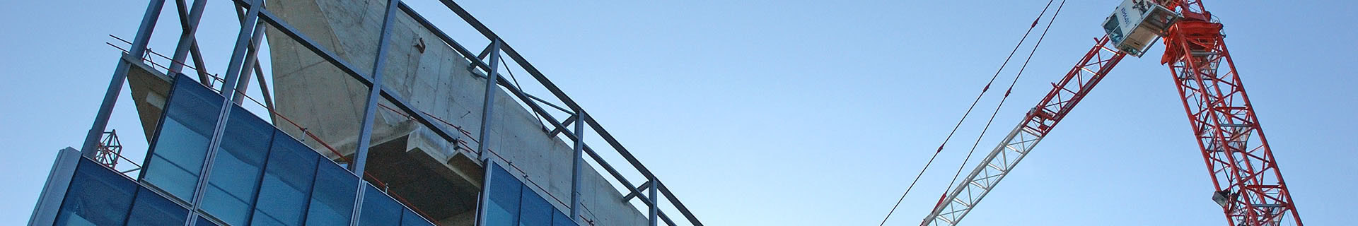 Das Bild zeigt eine Baustelle mit einem teilweise errichteten Gebäude und einem großen rot-weißen Baukran. Die unteren Stockwerke des Gebäudes sind bereits mit Glasfenstern versehen, die oberen Stockwerke sind noch im Rohbau mit sichtbarem Beton und Stahlgerüst. Der Kran ragt hoch über das Gebäude und deutet auf aktive Bauarbeiten hin. 
