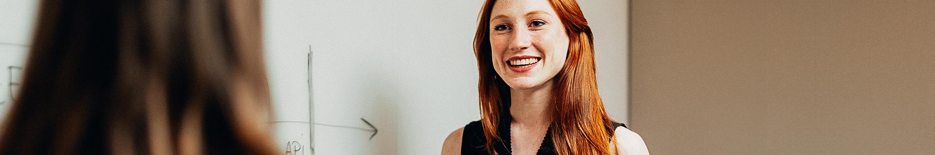 Das Foto zeigt eine Unterrichtssituation mit zwei Personen. Eine Person, deren Gesicht unscharf ist, blickt in die Kamera, während die andere von hinten zu sehen ist. Im Hintergrund befindet sich eine weiße Tafel mit einigen Schriftzeichen und einer einfachen Grafik mit einem nach rechts zeigenden Pfeil. Der Text auf der Tafel enthält „AP1“.